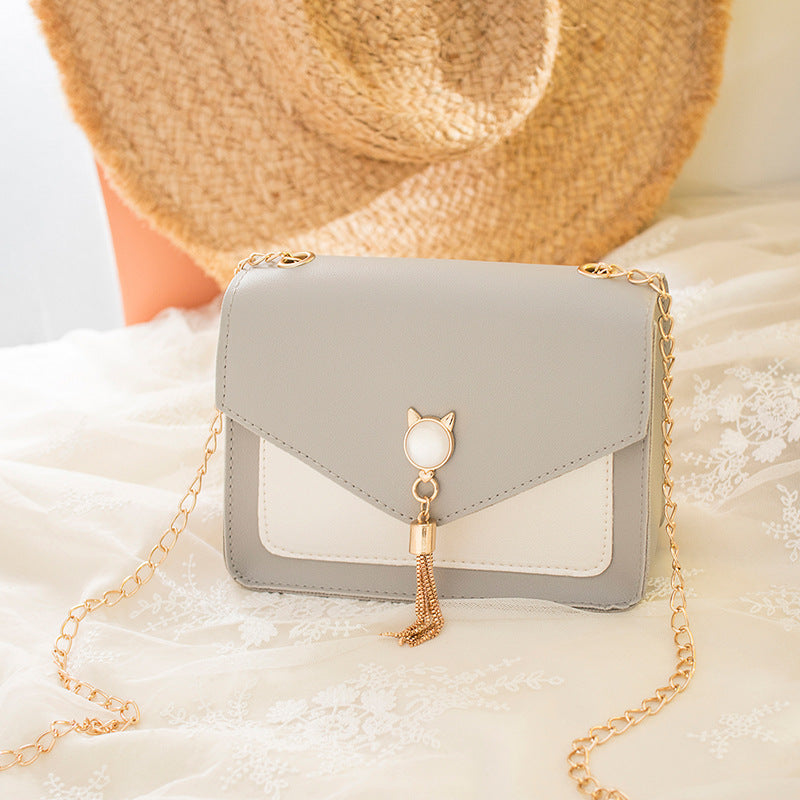 Gray handbag with gold chain and cat-shaped charm on a soft surface with a straw hat in the background