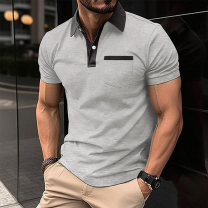 Man wearing a gray polo shirt with black collar and pocket trim, standing against a blurred background.